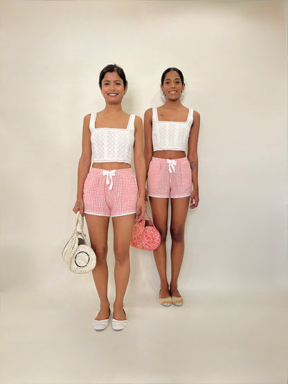 Two models wearing matching outfits with white embroidered corset tops and pink gingham shorts on a plain background