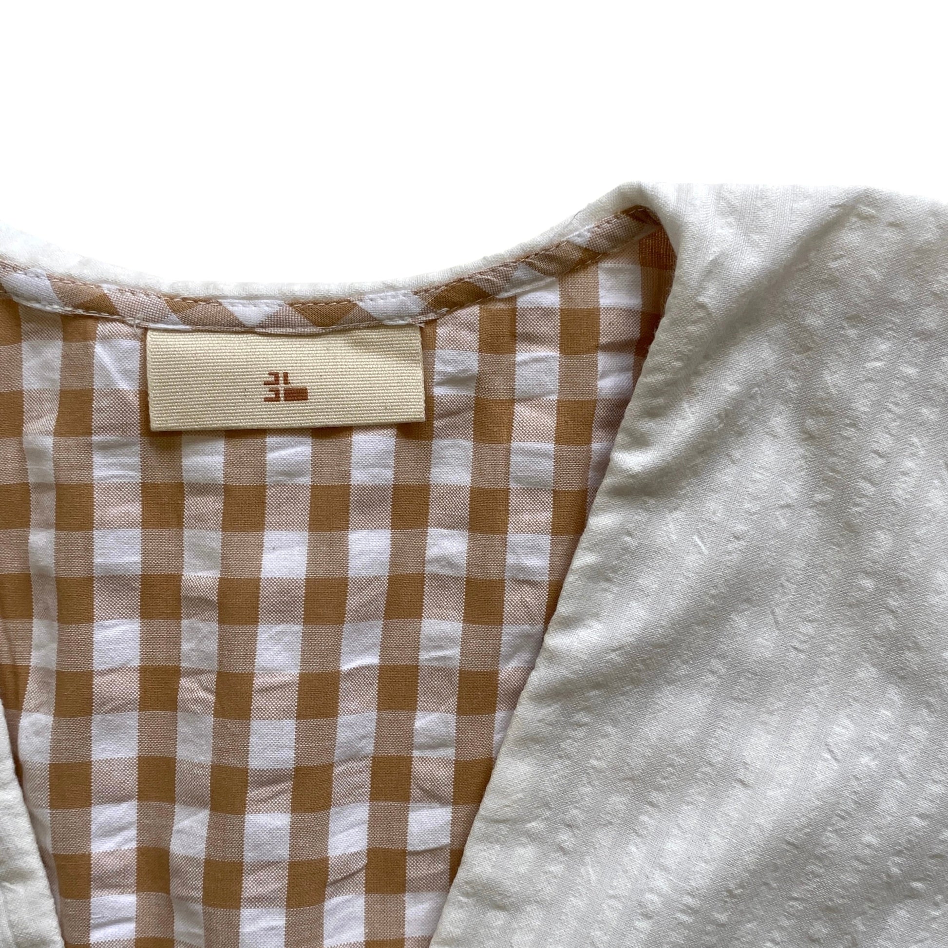 Close-up of a garment with a checkered pattern and a brand label on a white background. Organic cotton brown gingham blouse. White collar.