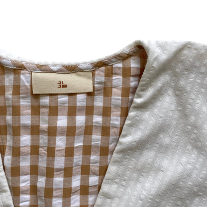Close-up of a garment with a checkered pattern and a brand label on a white background. Organic cotton brown gingham blouse. White collar.